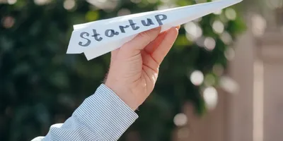 Hand hält Papierflieger mit dem Schriftzug 'Startup' vor unscharfem Hintergrund.