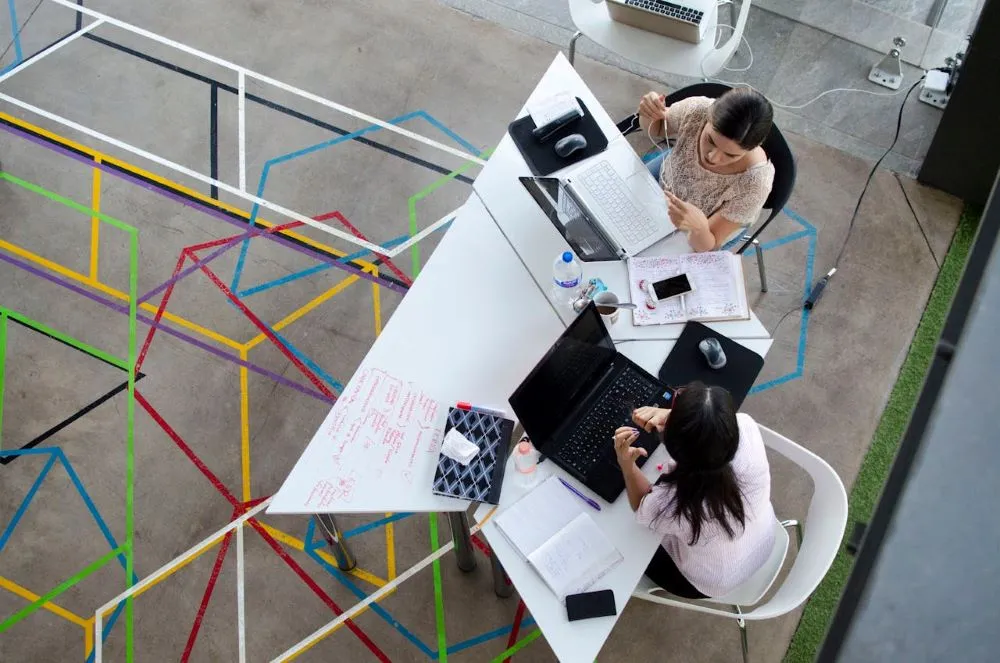 Zwei Frauen arbeiten an Laptops an einem weißen Tisch in einem modernen Büro mit farbigen Linien auf dem Boden