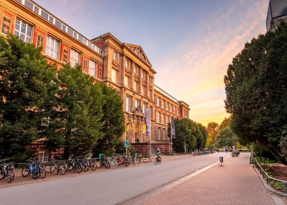 Historisches Gebäude der TU Darmstadt mit Fahrrädern davor und Bäumen an der Straße bei Sonnenuntergang.
