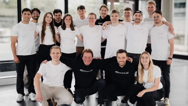 Gruppe von 13 Personen in weißen T-Shirts und einem schwarzen Hoodie, die in einem modernen Raum posieren. Einige Personen stehen, andere knien oder sitzen.