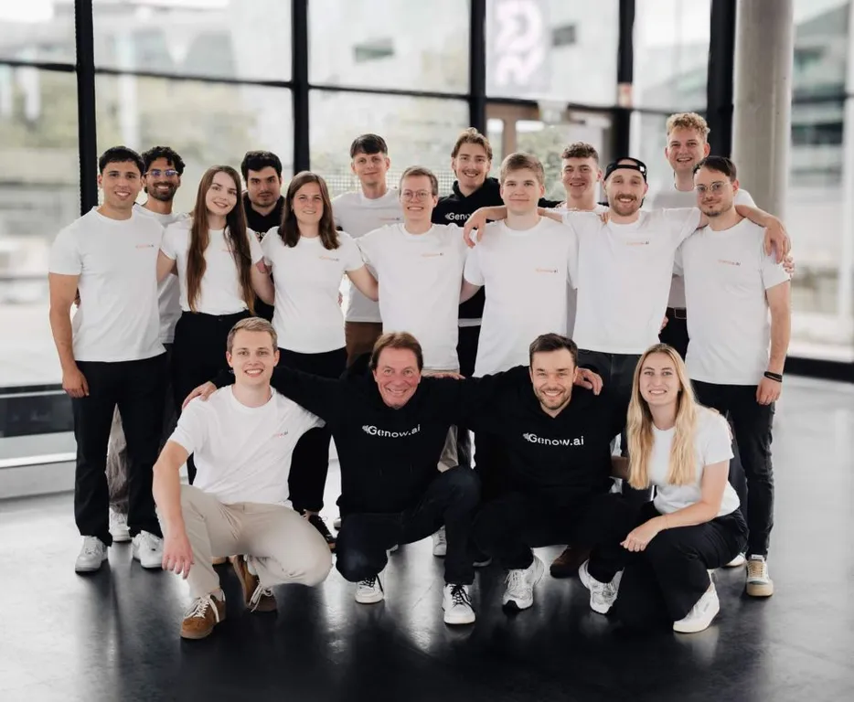 Gruppe von 13 Personen in weißen T-Shirts und einem schwarzen Hoodie, die in einem modernen Raum posieren. Einige Personen stehen, andere knien oder sitzen.