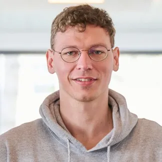 Person mit kurzen braunen Haaren trägt hellgrauen Kapuzenpullover vor unscharfem Hintergrund