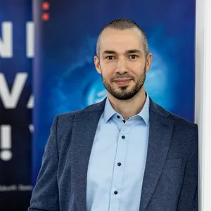 Tim Puppe, ein Mann mit dunklem Sakko und hellblauem Hemd vor blauem Hintergrund mit weißer und roter Schrift.