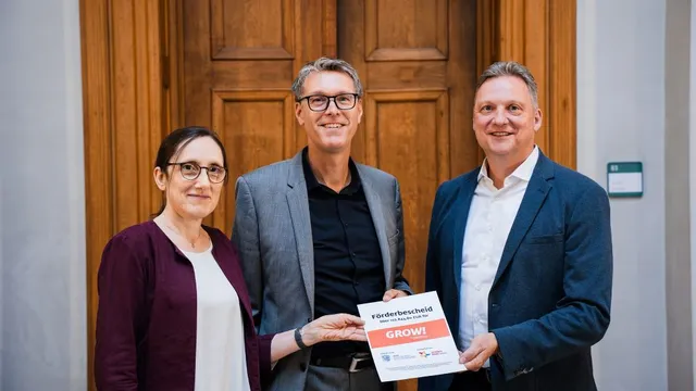 Drei Personen vor einer Holztür halten gemeinsam einen Förderbescheid mit der Aufschrift 'GROW!'