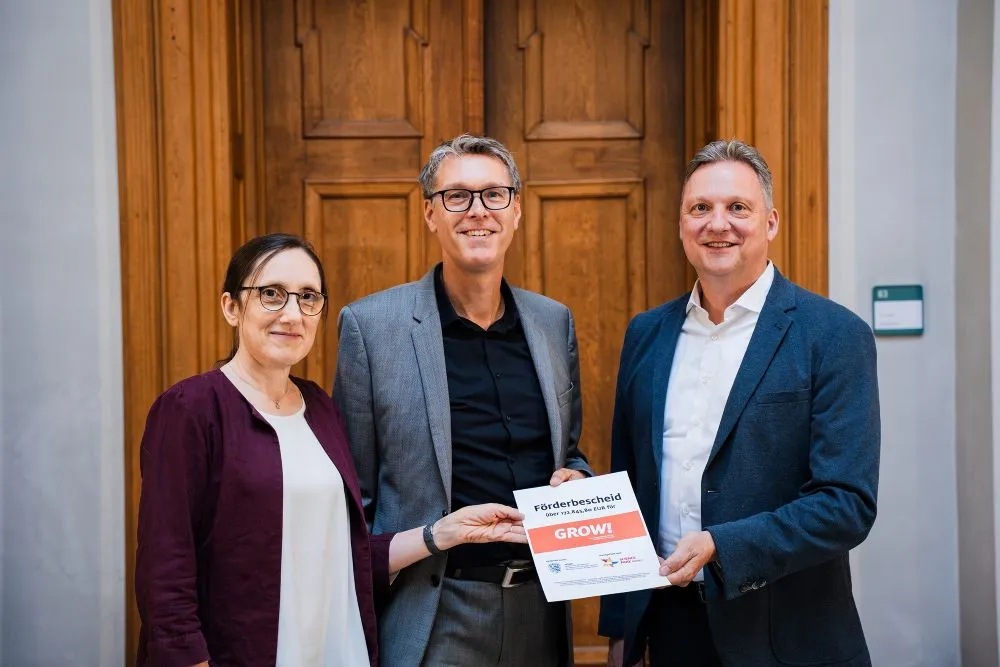 Drei Personen vor einer Holztür halten gemeinsam einen Förderbescheid mit der Aufschrift 'GROW!'