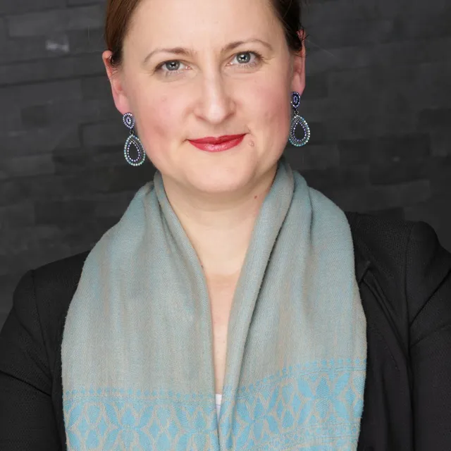 Oberkörper einer Person mit hochgestecktem Haar, die einen schwarzen Blazer mit silbernen Knöpfen, einen hellen Schal und auffällige Ohrringe trägt