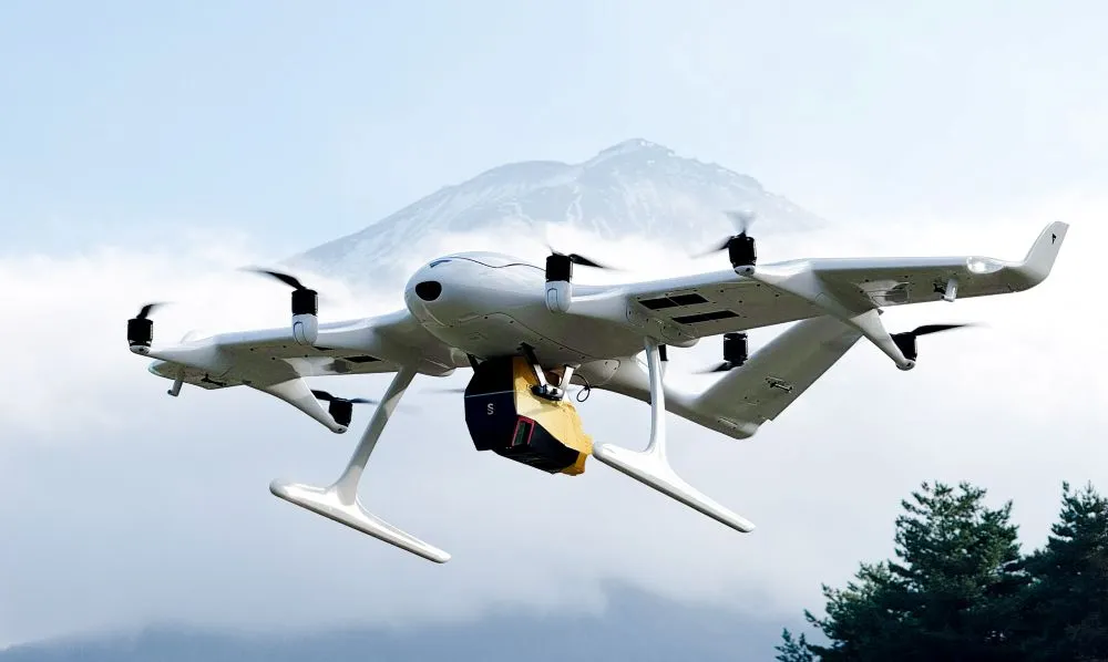 Drohne mit vier Rotoren und einem gelben Paket unter dem Rumpf vor bewölktem Himmel und Berg im Hintergrund