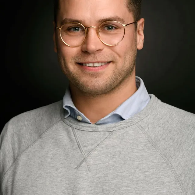 Oberkörper eines Mannes mit grauem Pullover über blauem Hemd vor schwarzem Hintergrund
