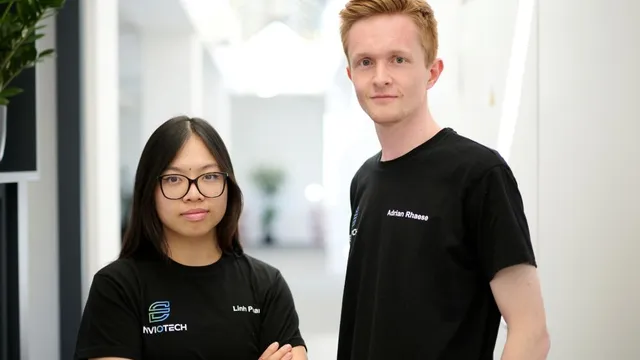 Zwei Personen stehen nebeneinander in einem hellen Flur, beide tragen schwarze T-Shirts mit weißen Logos und Namen, die Arme der linken Person sind verschränkt