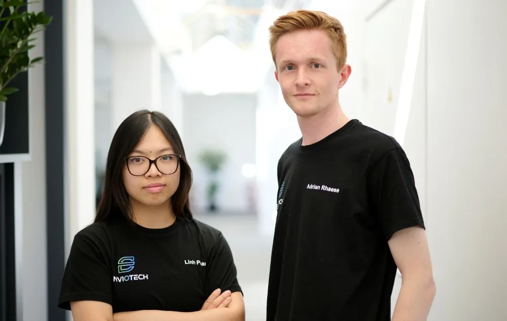 Zwei Personen stehen nebeneinander in einem hellen Flur, beide tragen schwarze T-Shirts mit weißen Logos und Namen, die Arme der linken Person sind verschränkt