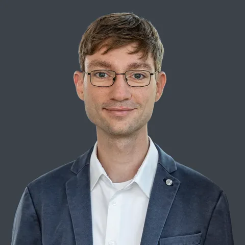 Porträt eines lächelnden Mannes mit Brille, weißem Hemd und dunkelblauem Sakko vor einfarbigem Hintergrund
