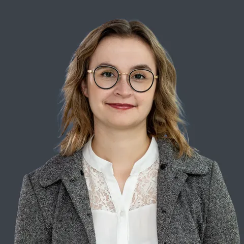 Frau mit schulterlangen gewellten Haaren, runder Brille, weißer Spitzenbluse und grauem Blazer vor einfarbigem Hintergrund