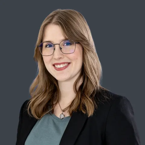 Lächelnde Frau mit schulterlangen blonden Haaren, Brille, grauem Oberteil und schwarzem Blazer vor einfarbigem Hintergrund