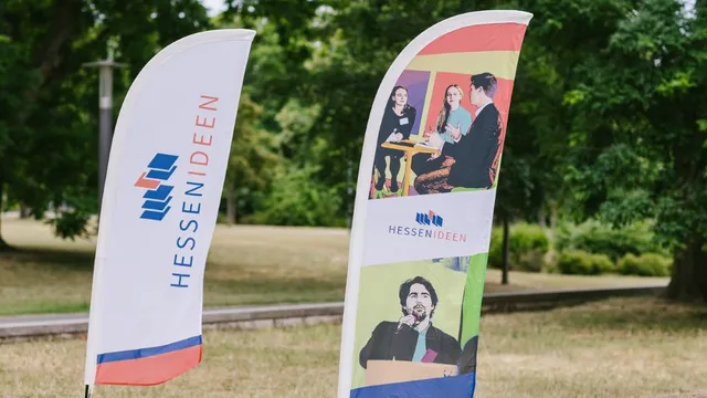 Zwei stehende Fahnen mit dem Logo und Schriftzug 'HESSEN IDEEN', eine zeigt Fotos von Personen in Gesprächssituationen, die andere ist weiß mit blau-rotem Rand.