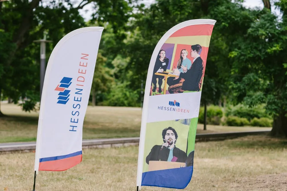 Zwei stehende Fahnen mit dem Logo und Schriftzug 'HESSEN IDEEN', eine zeigt Fotos von Personen in Gesprächssituationen, die andere ist weiß mit blau-rotem Rand.
