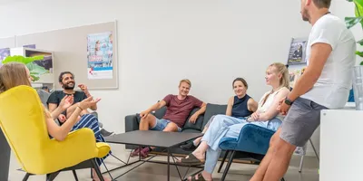 Mehrere Personen sitzen in einem Raum auf verschiedenen Stühlen und einem Sofa, eine Person lehnt an einem Tisch, im Hintergrund Poster an der Wand.