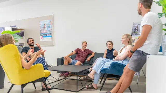 Mehrere Personen sitzen in einem Raum auf verschiedenen Stühlen und einem Sofa, eine Person lehnt an einem Tisch, im Hintergrund Poster an der Wand.