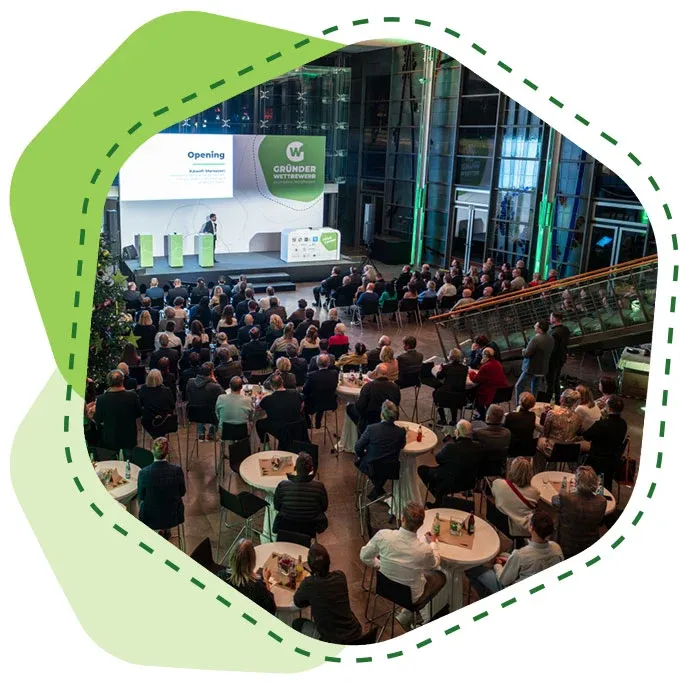 Publikum sitzt an runden Tischen in einem großen Saal und hört einer Person auf einer Bühne mit Präsentation zu.