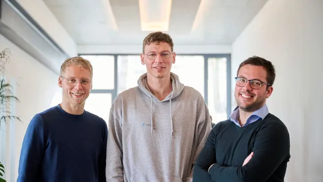 Drei Männer stehen nebeneinander in einem hellen Raum mit großen Fenstern, der mittlere trägt einen Kapuzenpullover, die anderen zwei Pullover und Hemd.