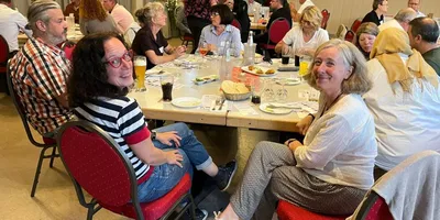 Eine Gruppe von Personen sitzt an einem langen Tisch in einem Restaurant. Auf dem Tisch sind Teller mit Essen, Gläser und Getränke sichtbar. Die Gäste sind in lockerer Kleidung und unterhalten sich.