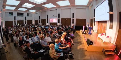 Großer Saal mit Publikum bei einer Präsentation vor Leinwand und Rednerpult