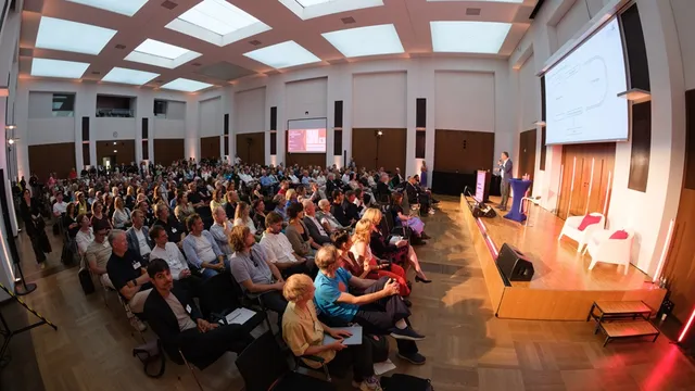 Großer Saal mit Publikum bei einer Präsentation vor Leinwand und Rednerpult