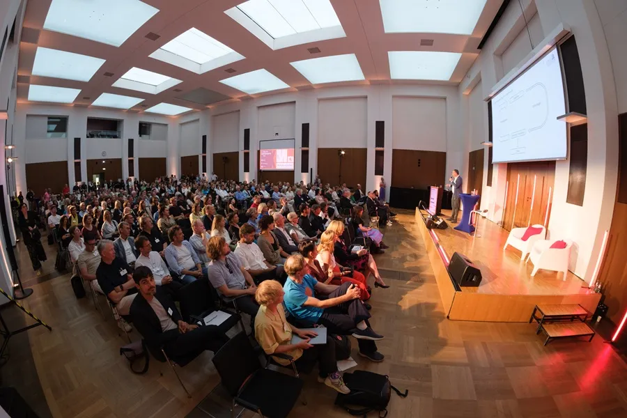 Großer Saal mit Publikum bei einer Präsentation vor Leinwand und Rednerpult
