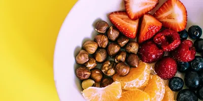 Weißer Teller mit Haselnüssen, Mandarinenstücken, Erdbeerhälften, Himbeeren und Blaubeeren vor gelbem Hintergrund.