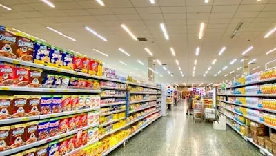 Supermarket aisle with shelves stocked with various cereal boxes on the left and other packaged goods on the right, with shoppers in the distance.