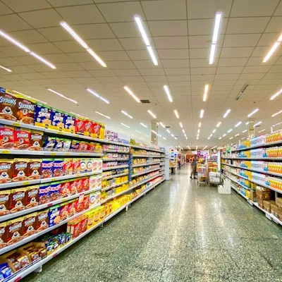 Supermarket aisle with shelves stocked with various cereal boxes on the left and other packaged goods on the right, with shoppers in the distance.