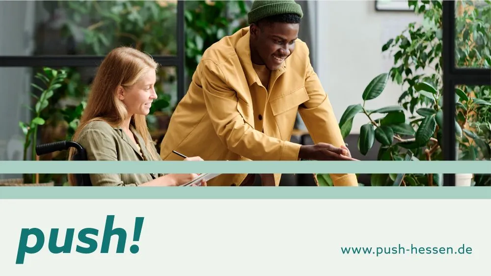 Zwei Personen sitzen an einem Tisch mit Pflanzen im Hintergrund, eine Person trägt eine grüne Mütze und eine gelbe Jacke, die andere Person hält einen Stift und ein Notizbuch.