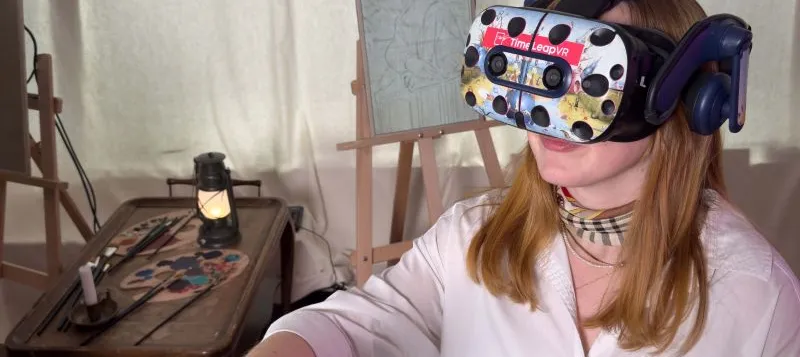 Person mit langen Haaren trägt eine VR-Brille und hält zwei VR-Controller in einem Raum mit Staffelei und Malutensilien