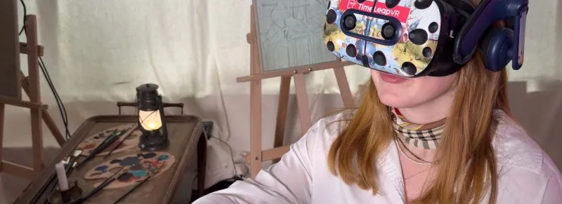 Person mit langen Haaren trägt eine VR-Brille und hält zwei VR-Controller in einem Raum mit Staffelei und Malutensilien